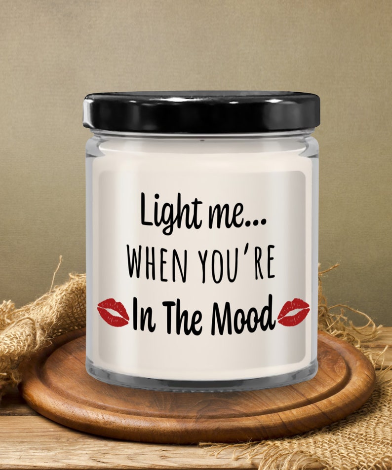 May include: A white candle in a glass jar with a black lid. The candle has the text "Light me... When you're In The Mood" with red lips on either side of the text.