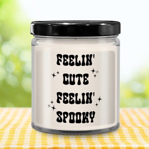 May include: A clear glass candle jar with a black lid, filled with white wax. The label features the text "FEELIN' CUTE FEELIN' SPOOKY" in black, with star accents. The candle sits on a yellow and white checkered tablecloth.