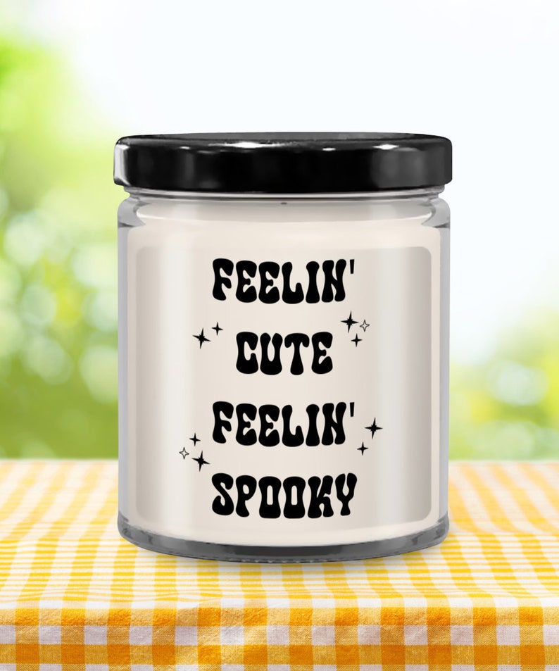 May include: A clear glass candle jar with a black lid, filled with white wax. The label features the text "FEELIN' CUTE FEELIN' SPOOKY" in black, with star accents. The candle sits on a yellow and white checkered tablecloth.