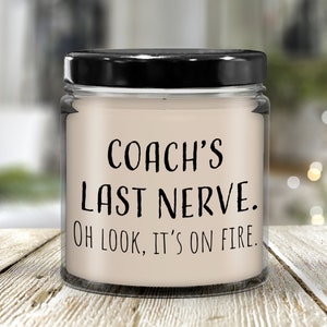 May include: A glass jar candle with a black lid. The candle is white and has the text "COACH'S LAST NERVE. OH LOOK, IT'S ON FIRE." printed on it.