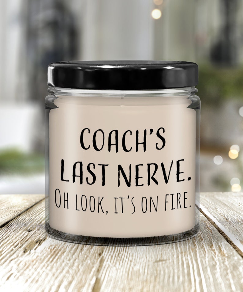 May include: A glass jar candle with a black lid. The candle is white and has the text "COACH'S LAST NERVE. OH LOOK, IT'S ON FIRE." printed on it.