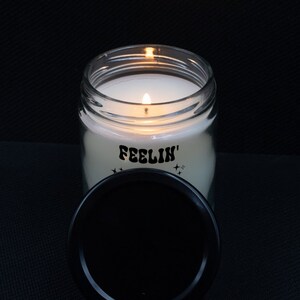 May include: A lit candle in a clear glass jar with a black lid. The candle has the word "FEELIN'" printed on the front with star graphics. The lid is off to the side.