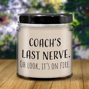 May include: A glass jar candle with a black lid and a white label. The label reads "Coach's Last Nerve. Oh look, it's on fire."