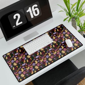 Puede incluir: Alfombrilla de escritorio rectangular con un estampado floral en tonos amarillos, rosas, morados y verdes sobre un fondo marrón oscuro. Un teclado y un ratón blancos están sobre la alfombrilla, con un monitor de ordenador al fondo.