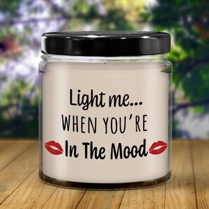 May include: A white candle in a clear glass jar with a black lid. The candle has the text "Light me... When you're In The Mood" with red lips on either side of the text.
