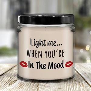 May include: A clear glass candle jar with a black lid. The candle is white and has the text "Light me... When you're In The Mood" with red lips on either side of the text.