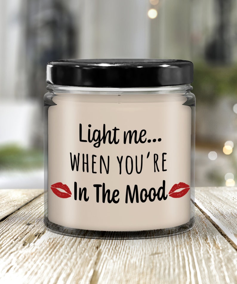 May include: A clear glass candle jar with a black lid. The candle is white and has the text "Light me... When you're In The Mood" with red lips on either side of the text.