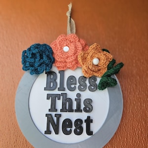 May include: A round wooden sign with a gray frame and the words "Bless This Nest" in black lettering. The sign is decorated with three crocheted flowers in shades of orange, teal, and brown.