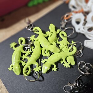 Könnte beinhalten: Eine Gruppe von sechs limegrünen Gecko-Schlüsselanhängern mit silbernen Schlüsselringen. Die Schlüsselanhänger liegen auf einer schwarzen Oberfläche.