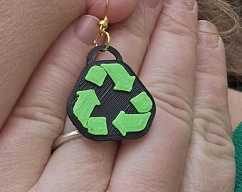 May include: A pair of black and green earrings shaped like a recycling symbol. The earrings are made of plastic and have a gold hook.