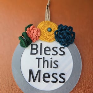 May include: A round gray sign with the text "Bless This Mess" in black lettering. The sign is decorated with crocheted flowers in yellow, green, blue, and orange.