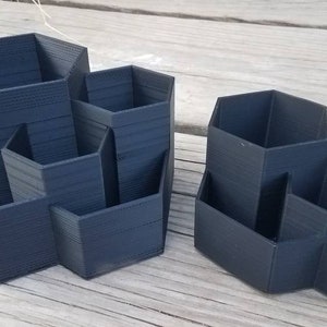 May include: Two black hexagonal desk organisers with multiple compartments. The organiser on the left has five compartments and the organiser on the right has four compartments.