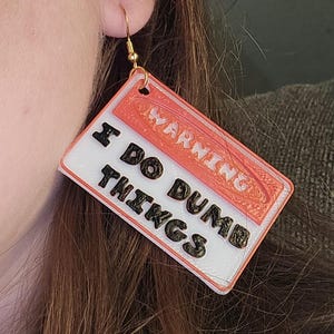 Puede incluir: Un par de pendientes con un letrero rojo y blanco que dice "WARNING I DO DUMB THINGS". El letrero está impreso en 3D y tiene un gancho dorado.