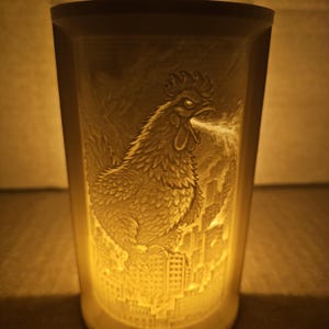 Puede incluir: Un portavelas cilíndrico de cerámica blanquecina con un relieve detallado de un gallo que respira fuego sobre un horizonte urbano. El interior está iluminado, creando un efecto cálido y brillante. El diseño es intrincado y artístico.