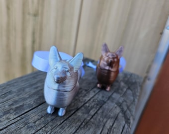 May include: Two 3D-printed corgi figurines, one silver and one bronze, each with a light blue ribbon. The corgis are standing on a weathered wooden surface. The figurines have detailed features.