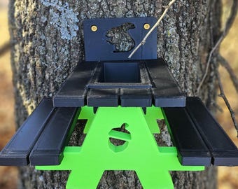 May include: A black and green squirrel feeder shaped like a picnic table with a squirrel silhouette on the back. The feeder is mounted on a tree trunk.