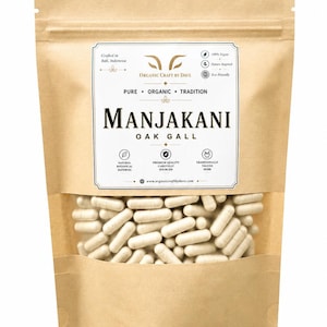 Può includere: Un sacchetto di carta marrone con una finestra trasparente che mostra numerose capsule beige. La borsa presenta un'etichetta bianca con il testo "MANJAKANI OAK GALL" e "ORGANIC CRAFT BY DAVE". Il testo aggiuntivo include "Pure, Organic, Tradition".