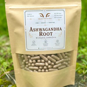 May include: A tan pouch with a clear window displaying numerous brown capsules. The pouch features the text "Ashwagandha Root" and "Withania somnifera" in a decorative label. Additional text includes "Organic Craft by Dave" and mentions of natural ingredients and traditional botanical material.