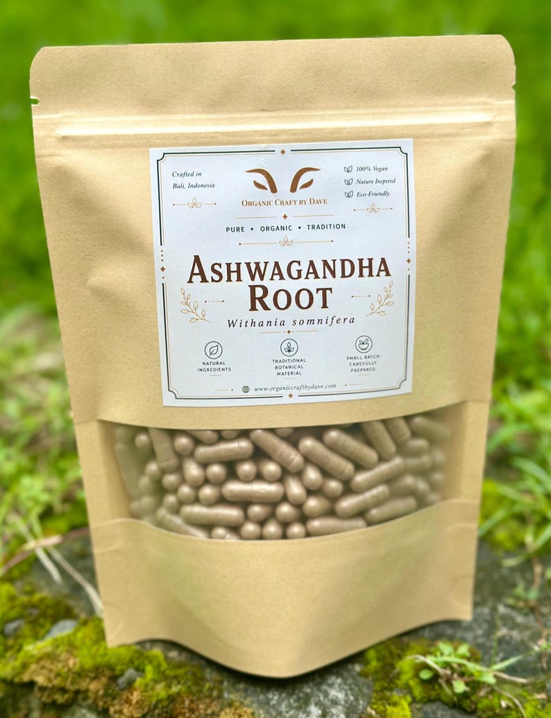 May include: A tan pouch with a clear window displaying numerous brown capsules. The pouch features the text "Ashwagandha Root" and "Withania somnifera" in a decorative label. Additional text includes "Organic Craft by Dave" and mentions of natural ingredients and traditional botanical material.