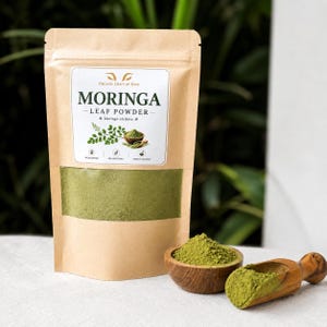 May include: A brown paper pouch containing green moringa leaf powder with a clear window. The label reads Organic Craft by Dave, Moringa Leaf Powder, Moringa oleifera, Wildcrafted, No Additives, Finely Ground. A wooden bowl and scoop hold the powder.