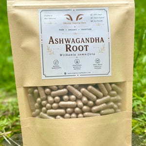 May include: A brown paper bag with a clear window displaying numerous beige capsules. The bag features a label with the text "Ashwagandha Root" and "Withania somnifera." Additional text includes "Organic Craft by Dave" and mentions natural ingredients.