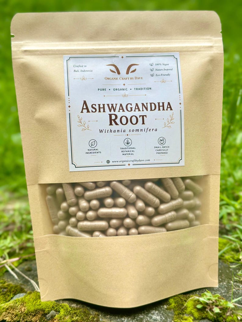 May include: A brown paper bag with a clear window displaying numerous beige capsules. The bag features a label with the text "Ashwagandha Root" and "Withania somnifera." Additional text includes "Organic Craft by Dave" and mentions natural ingredients.