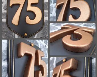 May include: Five different views of a house number plaque with the number 75 in gold raised lettering on a black background. The plaque is surrounded by a gold trim.