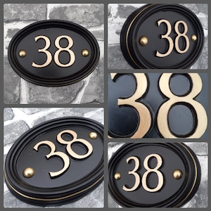 Op de afbeelding: Zwarte ovale huisnummerplaat met goudkleurig nummer "38" en gouden accenten.