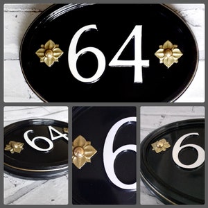 Puede incluir: Placa de número de casa ovalada negra con números blancos "64" y detalles decorativos dorados.