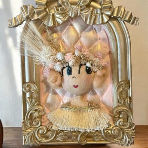 May include: A decorative doll in a gold frame. The doll has a painted face, a floral crown, and a gold tutu. The frame features ornate scrollwork and a bow at the top. The background is a quilted pink fabric.