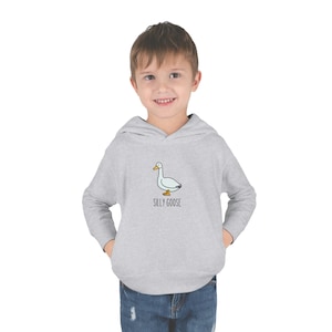 Puede incluir: Sudadera con capucha gris claro con un diseño de ganso de dibujos animados y el texto "SILLY GOOSE". El ganso es blanco y azul claro con pico y patas amarillas. La sudadera tiene un bolsillo delantero. El niño lleva vaqueros.