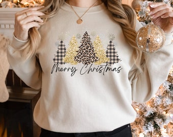 May include: A cream-colored sweatshirt with a festive design featuring three Christmas trees. The trees are decorated with gold glitter, black and white plaid, and leopard print. The text "Merry Christmas" is printed below the trees.