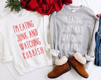 May include: Two white sweatshirts with the text "I'm eating junk and watching rubbish" in red on one and white on the other. The sweatshirts are laid out on a white surface with a pair of brown suede boots with white fur trim.