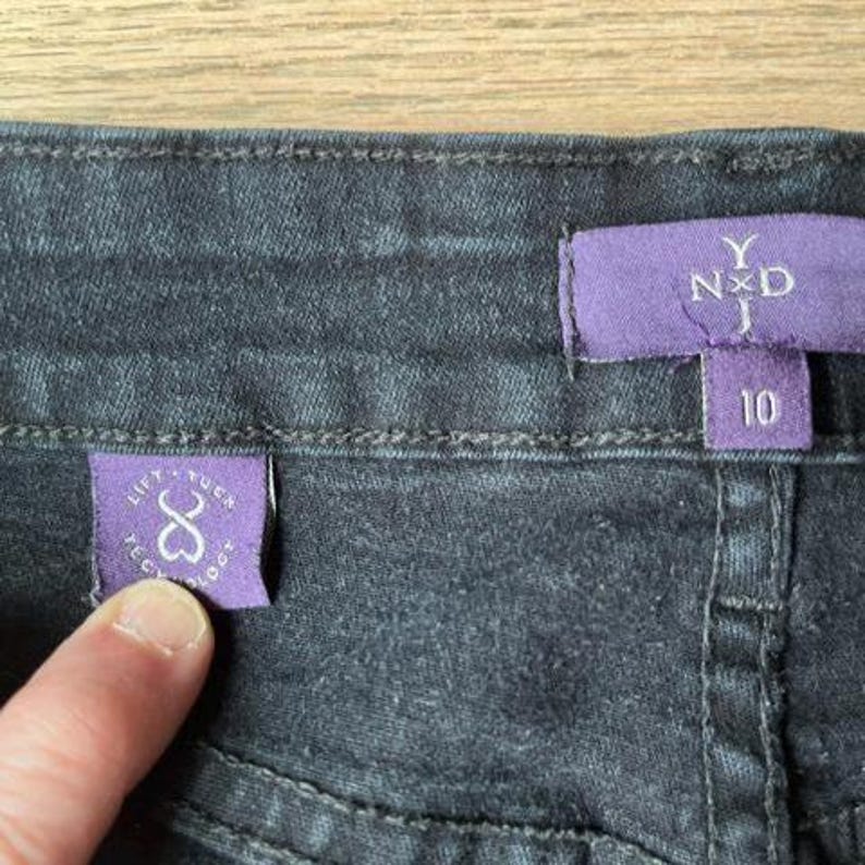 May include: Close-up of dark denim jeans with two purple tags. One tag reads "LIFT. TUCK. TECHNOLOGY" with a heart-shaped design. The other tag displays "Y NXD" and the number "10".