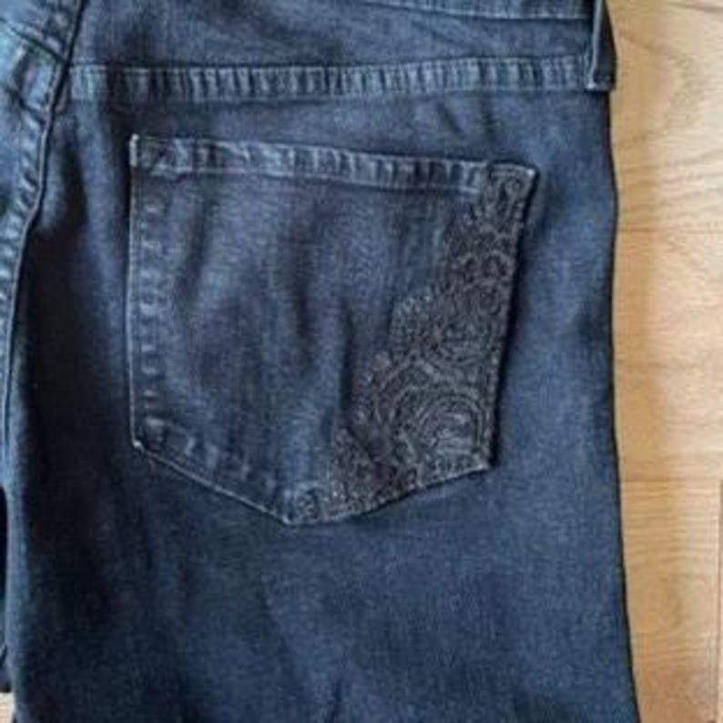 May include: Close-up of dark wash denim jeans. The jeans feature a decorative embroidered design on the back pocket. The denim has a slightly faded appearance, with visible stitching and a classic five-pocket design.