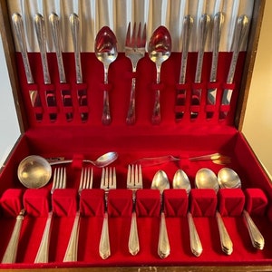 May include: A vintage silverware set in a wooden case with a red velvet interior. The set includes spoons, forks, and knives, neatly arranged. The case is open, revealing the polished silver utensils. The silverware is displayed against a white satin backdrop.