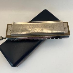 May include: A silver harmonica with the words "Professional Hohner Ultra-Monica No. 12" engraved on the top. The harmonica has a black case.