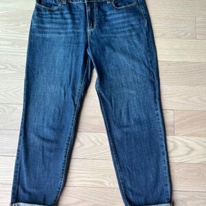 May include: Blue denim jeans with a classic five-pocket design. The jeans have a straight leg cut with cuffed hems. The wash is a medium blue with subtle fading and whiskering details. The jeans are laid flat on a light wood surface.