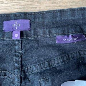 May include: Close-up of dark denim jeans with a straight leg cut. The jeans have a purple label with the brand name "Y N D J" and size "10". Another purple label reads "STRAIGHT". The jeans are made of a dark wash denim.
