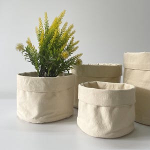 May include: Four off-white canvas storage baskets, one with a green plant inside. The baskets are made of a natural fabric and have a simple, rustic design.