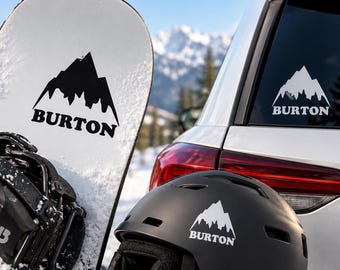 May include: A white snowboard with black bindings and a black mountain logo with the text BURTON, a black helmet with a white mountain logo and the text BURTON, and a white car window featuring a white mountain decal with the text BURTON.