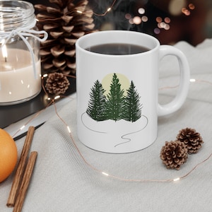 May include: White ceramic mug with a black handle. The mug has a graphic of three green pine trees with a yellow sun behind them. The trees are on a white background with a black line drawing of a path.