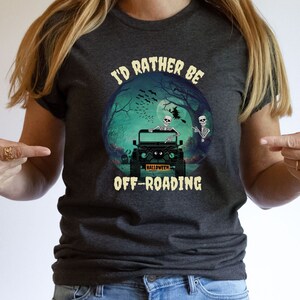 May include: A dark gray t-shirt with the text "I'd rather be off-roading" in white. A circle graphic features two skeletons in a jeep with a "Halloween" license plate. The background is a dark green forest with bats flying around a full moon.