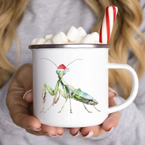 May include: White enamel mug with a green praying mantis wearing a red Santa hat. The mug has a silver rim and handle. There are white marshmallows in the mug.