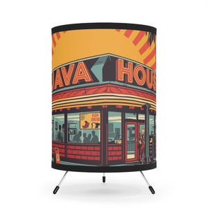 May include: A cylindrical lamp with a retro illustration of a coffee shop, "JAVA HOUSE", in red, teal, and yellow. The lamp has a black base and three metal legs. The design features a palm tree and a sign that says "Black Java".