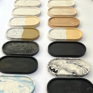 Peut inclure: Une collection de 14 plateaux ovales avec différents motifs et couleurs. Les plateaux sont faits d'un matériau moucheté, marbré ou uni. Les plateaux sont disposés en rangée sur une surface blanche.