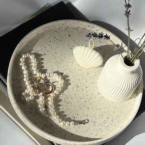 Op de afbeelding: Een witte ronde dienblad met een gespikkeld patroon, met een parelketting, een witte schelp en een witte vaas met lavendelstengels.