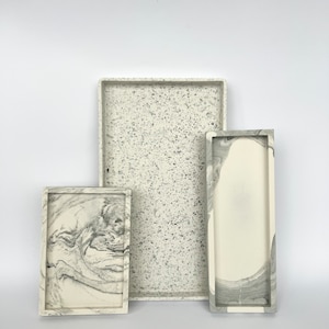 Peut inclure: Trois plateaux rectangulaires en résine blanche mouchetée et marbrée. Les plateaux sont de tailles différentes et sont empilés les uns sur les autres.