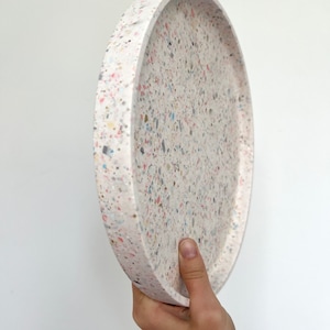 Könnte beinhalten: Ein runder, cremefarbener Tablett mit einem gesprenkelten Terrazzo-Design. Das Tablett wird schräg gehalten, wodurch der Rand und die farbenfrohen Flecken aus Blau, Rosa, Gelb und Grau auf der Oberfläche zur Geltung kommen. Das Tablett ist ein dekorativer Gegenstand.