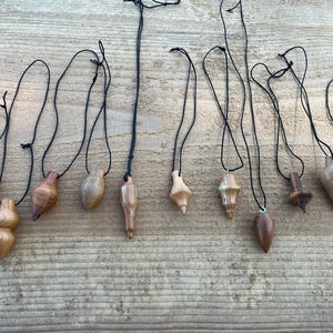 Könnte beinhalten: Eine Sammlung von Holzpendeln in verschiedenen Formen, darunter tropfenförmige, bauchige und konische Designs. Jedes Pendel ist an einer schwarzen Schnur befestigt. Die Pendel sind aus verschiedenen Holzarten gefertigt und zeigen eine Reihe natürlicher Farben und Maserungen.